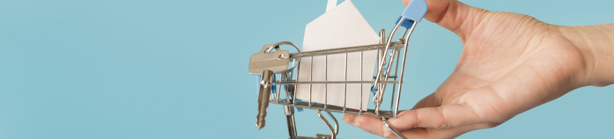 close-up-hand-holding-miniature-shopping-cart-with-paper-house-keys-against-blue-background