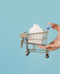 close-up-hand-holding-miniature-shopping-cart-with-paper-house-keys-against-blue-background