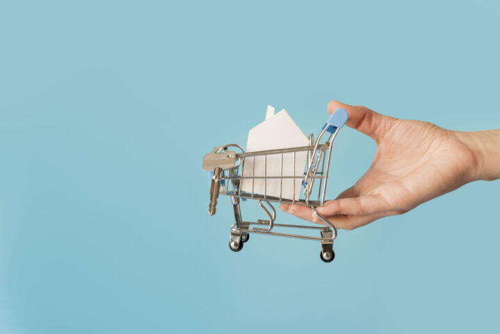 close-up-hand-holding-miniature-shopping-cart-with-paper-house-keys-against-blue-background