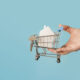 close-up-hand-holding-miniature-shopping-cart-with-paper-house-keys-against-blue-background