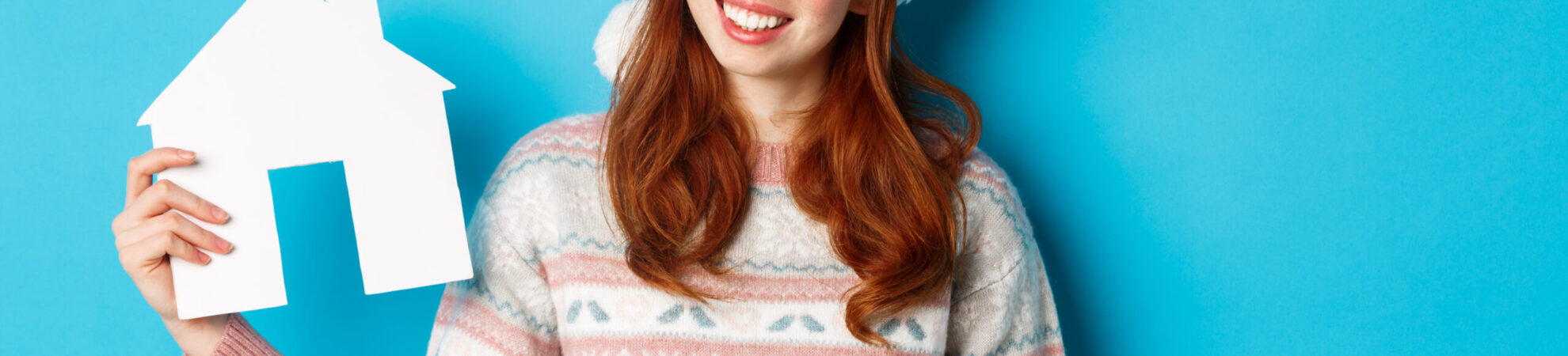 Holiday promos and real estate concept. Cheerful redhead woman in santa hat holding paper house in hand and smiling, standing in sweater against blue background.