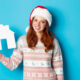 Holiday promos and real estate concept. Cheerful redhead woman in santa hat holding paper house in hand and smiling, standing in sweater against blue background.