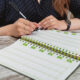 Women hands with pen write in notebook. Young girl at table in cozy office and fills out diary. Close-up of hands taking notes in magazine. Female works in an office. Planning of working time